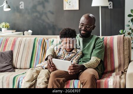 Happy African father sitting on sofa and playing online game on digital tablet together with his son at home Stock Photo
