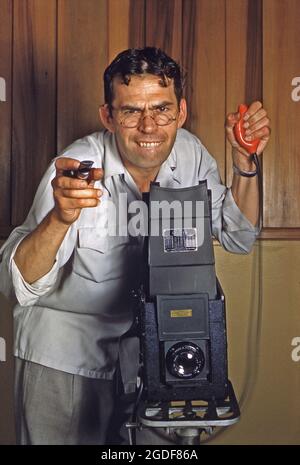 Studio photography in America in the 1950s – a photographer concentrates hard and is pressing a bulb shutter release with his huge Graflex camera. The bulb (a detachable rubber pneumatic device on a wire connected to the camera) meant the operator could help avoid camera shake, plus holding down the bulb and keeping the lens open was very useful for long time exposures. Graflex was an American camera manufacturer. This image is from an old American Kodak amateur colour transparency – a vintage 1950s photograph. Stock Photo