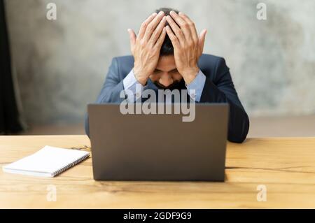 Stressed young Indian businessman touching head and feels dizzy, suffering from headache and migraine pain, sits at the desk with eyes closed, tired and exhausted man in formal suit has troubles Stock Photo