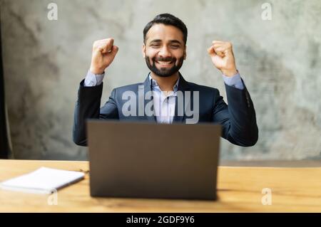 Excited and happy Indian businessman celebrating good deal or win sitting at desk, looks at the laptop screen, male entrepreneur in formal suit raising fists in triumph gesture, business going well Stock Photo