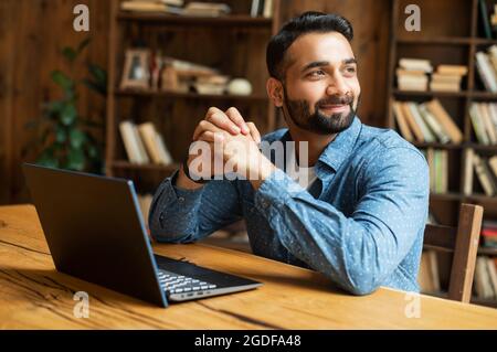 Happy and optimistic Indian man in casual wear using laptop in modern office, successful inspired arabic male entrepreneur looks away and smiles thoughtfully, planning next step Stock Photo