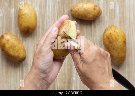 Top view of woman hands scrubbing boiled potato in jacket using knife on wooden background Stock Photo