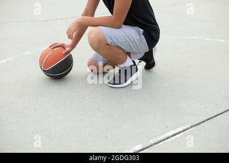 Male teenage basketball player crouching with ball on basketball court, neck down Stock Photo