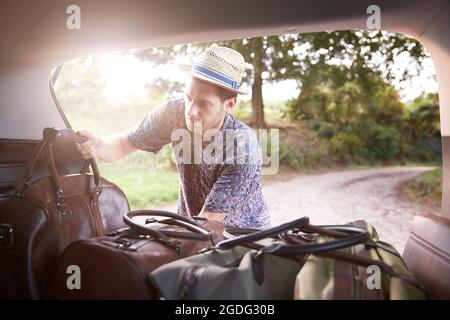 Young man in trilby removing luggage from car boot on rural road Stock Photo