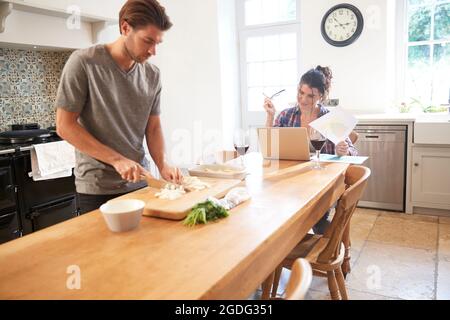 Man preparing vegetables at kitchen table, girlfriend using laptop Stock Photo
