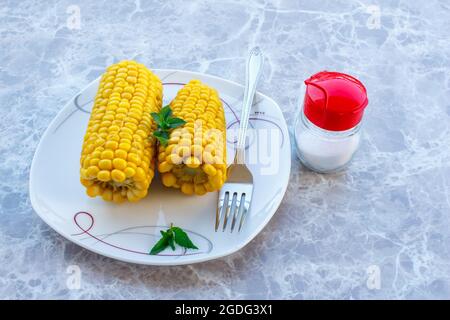 Tasty boiled corn cobs in plate with mint leaves and fork, on marble table background. Stock Photo