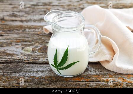 Glass jug of hemp milk with cannabis leaf on rustic table Stock Photo