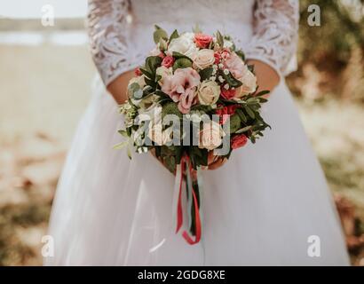 A bride in a white wedding dress holds a bouquet of red and white roses flowers in front of her outdoor Stock Photo