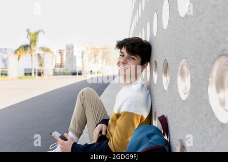 Happy young teen male sitting on urban bench while looking to camera Stock Photo