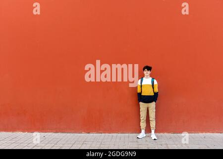 Young male with backpack standing on red wall while looking to camera Stock Photo