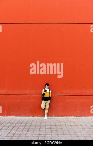 Young male with backpack standing on red wall using a mobile phone Stock Photo