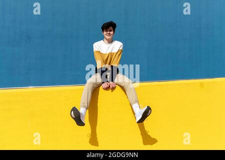 Confident man looking to camera while sitting on yellow retaining wall Stock Photo