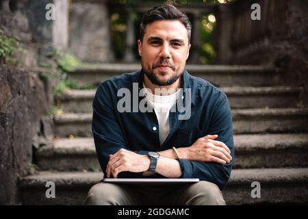 Portrait of happy man with laptop sitting on steps in city Stock Photo