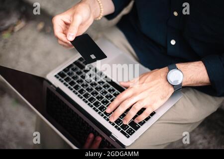 Man holding credit card while using laptop computer doing online shopping outdoors Stock Photo