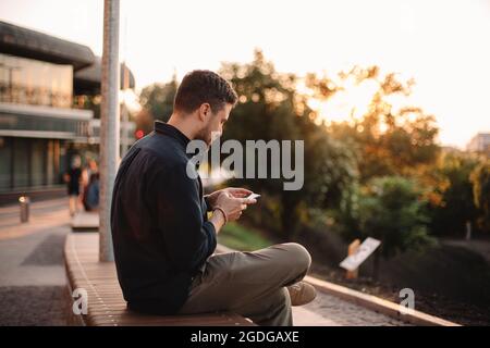 Happy man using smart phone sitting on bench at sunset Stock Photo