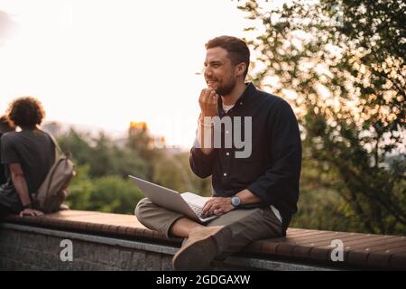 Happy thoughtful man using laptop computer sitting on bench in park in summer Stock Photo