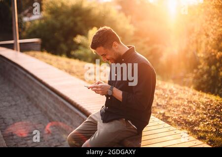 Happy man using smart phone sitting on bench at sunset Stock Photo