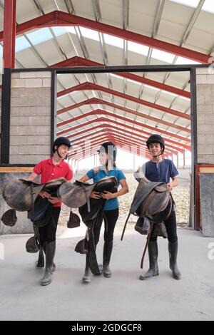 Mother and two sons holding horse riding saddles, smiling and talking to each other. Stock Photo