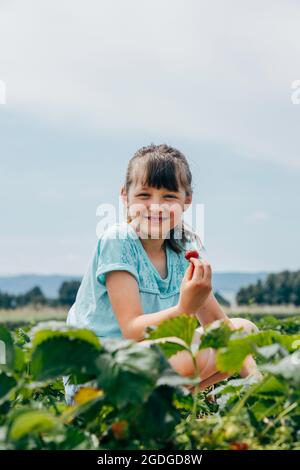 Smiling girl sitting in a strawberry field and holding strawberries in her hands against a blue sky. Stock Photo