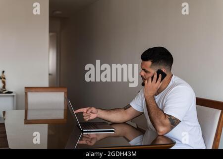 Tattooed Latino man touching laptop screen while talking on cell phone Stock Photo