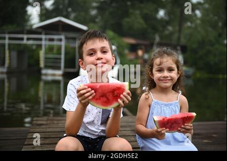 Children, kids, boy and girl sitting close to each other with slice of delicious juicy watermelon in hands and cute smiling to camera on the river bac Stock Photo