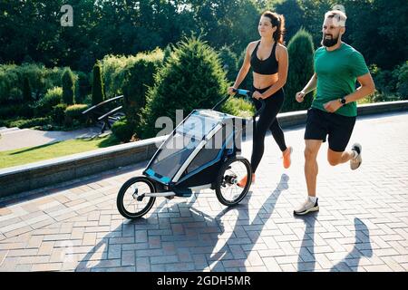 running couple with child in jogging stroller in public park at sunrise. Morning run for a whole family Stock Photo
