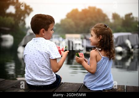 View from the back of two children, preschool girl and school boy, eating watermelon and looking at each other, enjoying beautiful sunset sitting on t Stock Photo