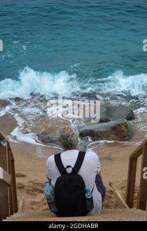 mature man with grey hair and a backpack sitting on a wooden ladder by the seaside Stock Photo