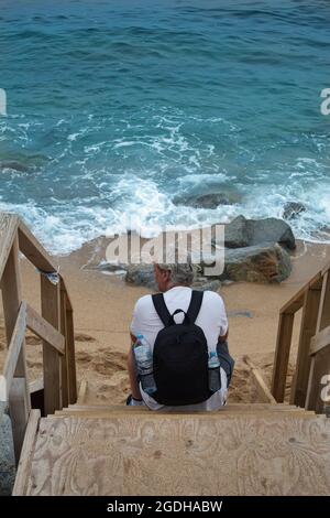 mature man with grey hair and a backpack sitting on a wooden ladder by the seaside Stock Photo