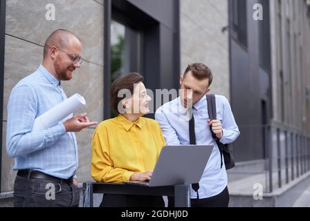 Designers or architect team looking at project blueprint and laptop as presentation outdoors. Business persons, men and woman in casual clothes standing outside, brainstorming project Stock Photo
