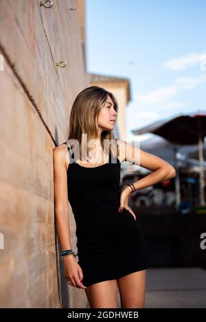Young blonde Caucasian girl with a black dress leaning against a wall in an uban setting Stock Photo