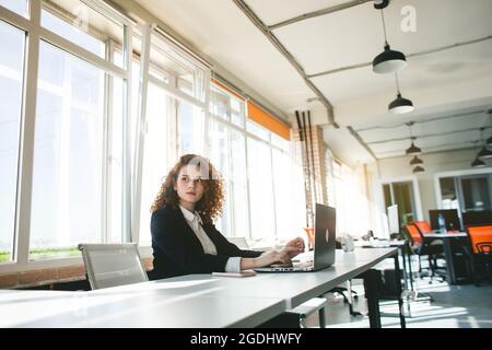Beautiful young red-haired woman in a business suit works in an office with a laptop Stock Photo