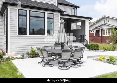 The backyard of a beautiful grey, luxury house with black accents, a covered seating area, large concrete patio, and landscaping. Stock Photo