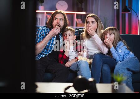Caucasian family of four sitting together on couch and eating popcorn while watching fascinating movie. Human feelings during interesting TV show. Stock Photo