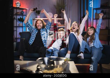 Emotional family of four celebrating winning in video games during free time at home. Happy parents with kids sitting on couch and taking fun together. Stock Photo