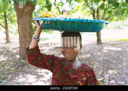 Grinning beautiful black girl carrying a big blue plastic plate on her head full of bananas Stock Photo