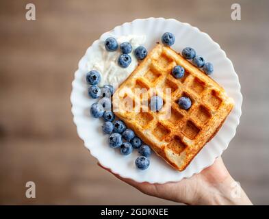A woman holds in her hand a plate on which fresh waffles with berries are ready. Homemade waffles with strawberries and blueberries. Delicious Breakfa Stock Photo