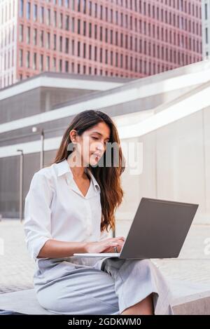 vertical portrait of an elegant young indian woman. She is sitting near tribunal building and using a laptop Stock Photo