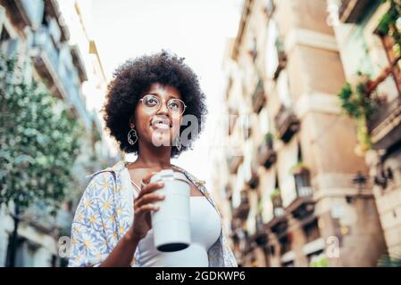 horizontal portrait of a young african woman. She is smiling and holds a reusable cup of coffee. Concept of summer morning in the city Stock Photo