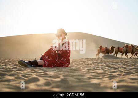 asian woman in red dress sitting in desert looking at view with caravan of camels and huge sand dune in background Stock Photo