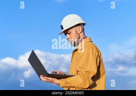 Building contractor, supervisor, instructor or construction engineer in white helmet and orange jacket using laptop at construction site. Sunny day with blue cloudy sky. High quality image Stock Photo
