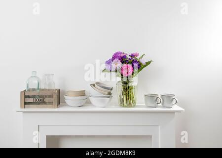 kitchen utensils, cups, bowls, bottles, storage items, a bouquet of asters are on a white dresser Stock Photo