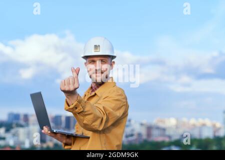 Bearded supervisor, foreman or construction inspector looking at camera and making Korean heart gesture. Contractor in hardhat and orange jacket holding computer laptop in front of sunny day blue sky Stock Photo