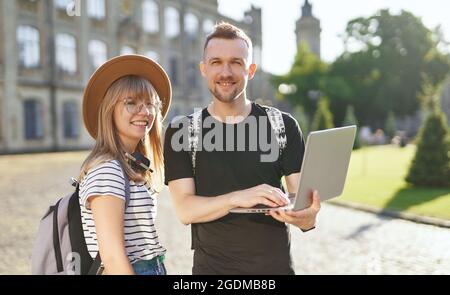Studying together. Two cute students, male and female, with backpacks using laptop at the university campus. Adorable successful students couple smiling looking at camera smiling. High quality photo Stock Photo