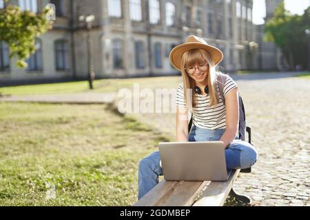 Concept of e-learning, distance study or remote learning concept, Young happy blonde school girl, college or university student with backpack and hat using laptop sitting on bench in university campus Stock Photo