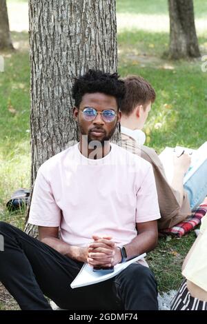 Portrait of African college student in eyeglasses sitting on the grass and looking at camera during studying outdoors Stock Photo
