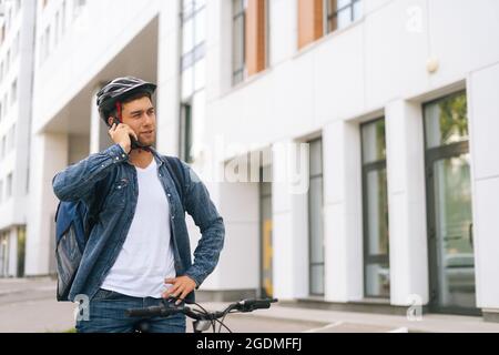 Medium shot of handsome courier male with thermal backpack talking on mobile phone to customer, looking for address to deliver online order. Stock Photo