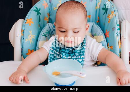Serious baby sits at baby chair and looking to empty plate. Baby waiting for food in his high chair