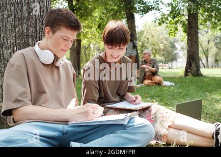 Two students sitting on the grass and making notes in notebooks they studying and working in team outdoors Stock Photo