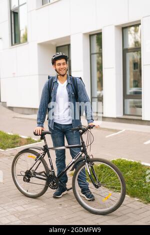 Full length vertical portrait of happy handsome delivery man in protective helmet posing standing near bicycle in city street, looking away Stock Photo